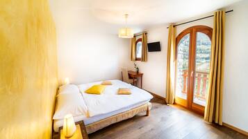 Quarto com cama, luminária de cabeceira, piso de madeira e vista para uma varanda com guarda-corpo de madeira.