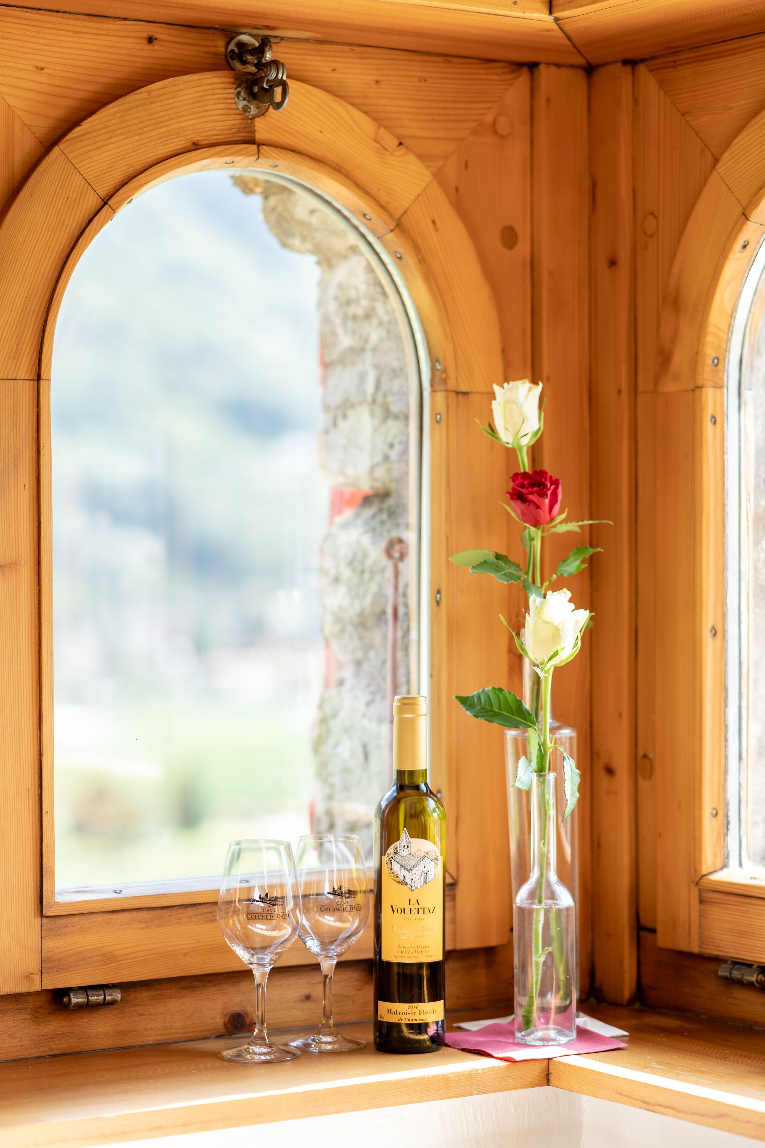 Een houten interieur met een groot boogvenster, een fles wijn, twee glazen en een vaas met bloemen.