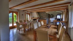 A dining area with a wooden table and chairs, a stone fireplace, and a view of greenery through large windows. - Castel de Daval (Sierre)