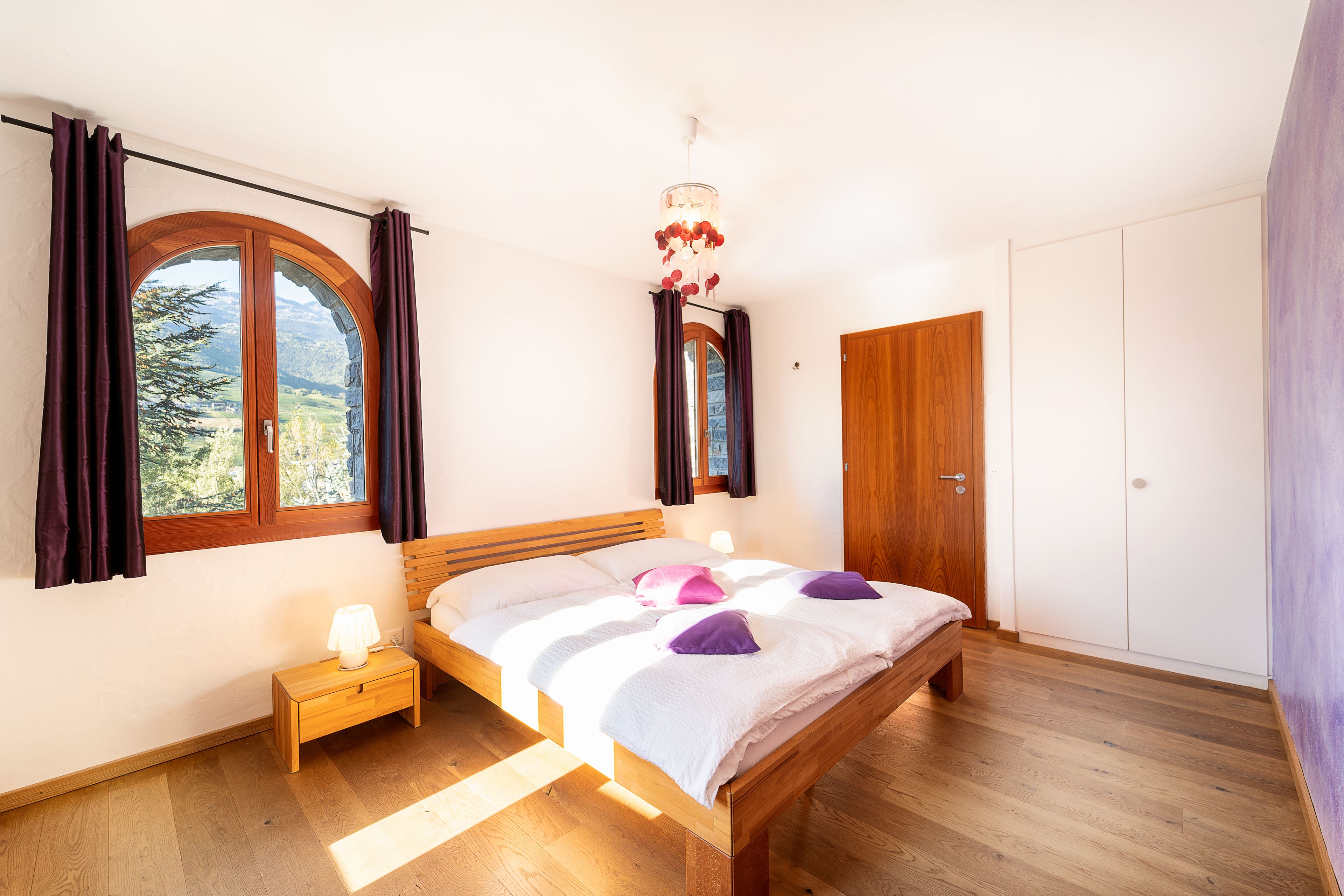 A bedroom with a wooden bed, a nightstand, a lamp, and a view of the outdoors through a window.