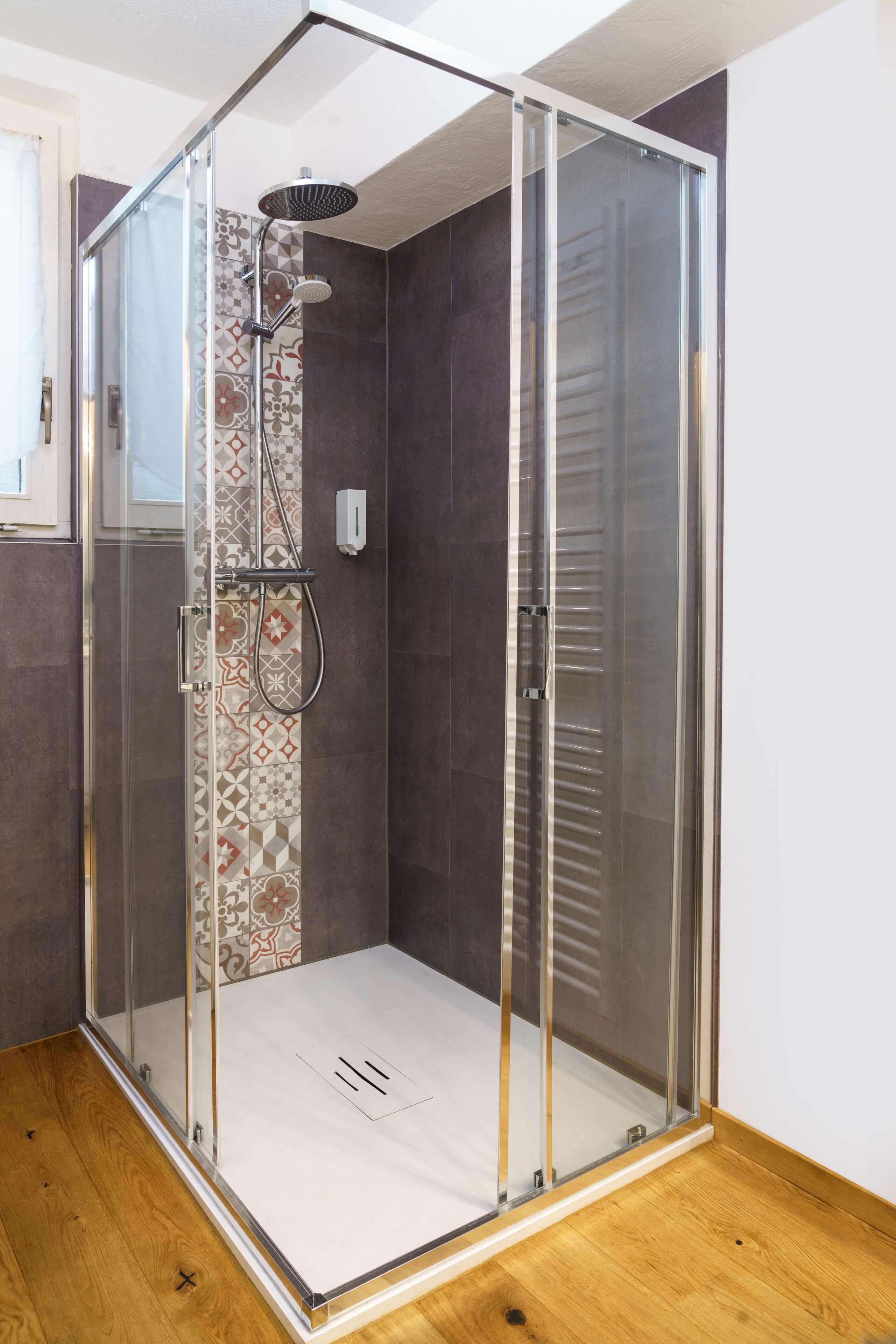 A glass-enclosed shower with a patterned tile wall, a wooden floor, and a white ceiling.