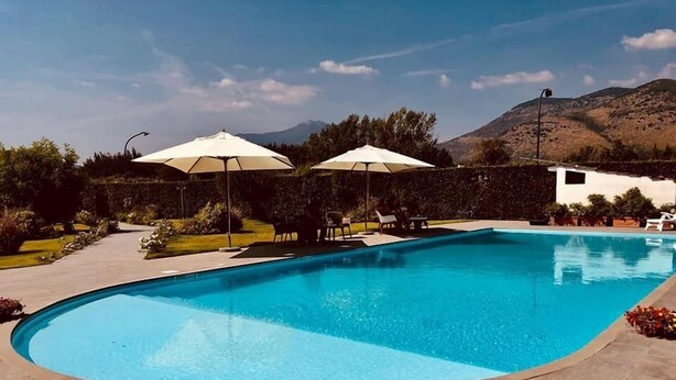Une piscine rectangulaire à bord incurvé, entourée d’une terrasse pavée. Deux parasols blancs offrent de l’ombre, et la piscine est située devant un paysage de collines et d’arbres.