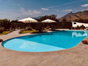A rectangular swimming pool with a curved edge, surrounded by a paved patio. There are two white umbrellas providing shade, and the pool is set against a backdrop of hills and trees. - Onorati Hotel (Priverno)