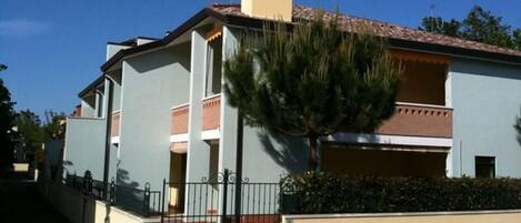 Un edificio de dos pisos con fachada azul claro, techo de tejas rojas y una chimenea central. Se observa una reja metálica en la parte delantera, y un árbol a la derecha.