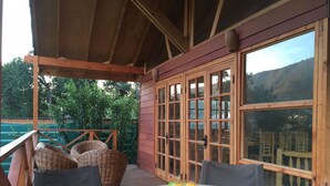 Una terraza de madera con sillas de mimbre y una mesa, un ventanal con puertas de cristal y vistas al verdor.