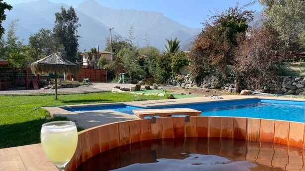 Una bañera de hidromasaje con borde de madera, un vaso de bebida transparente y una piscina al fondo.