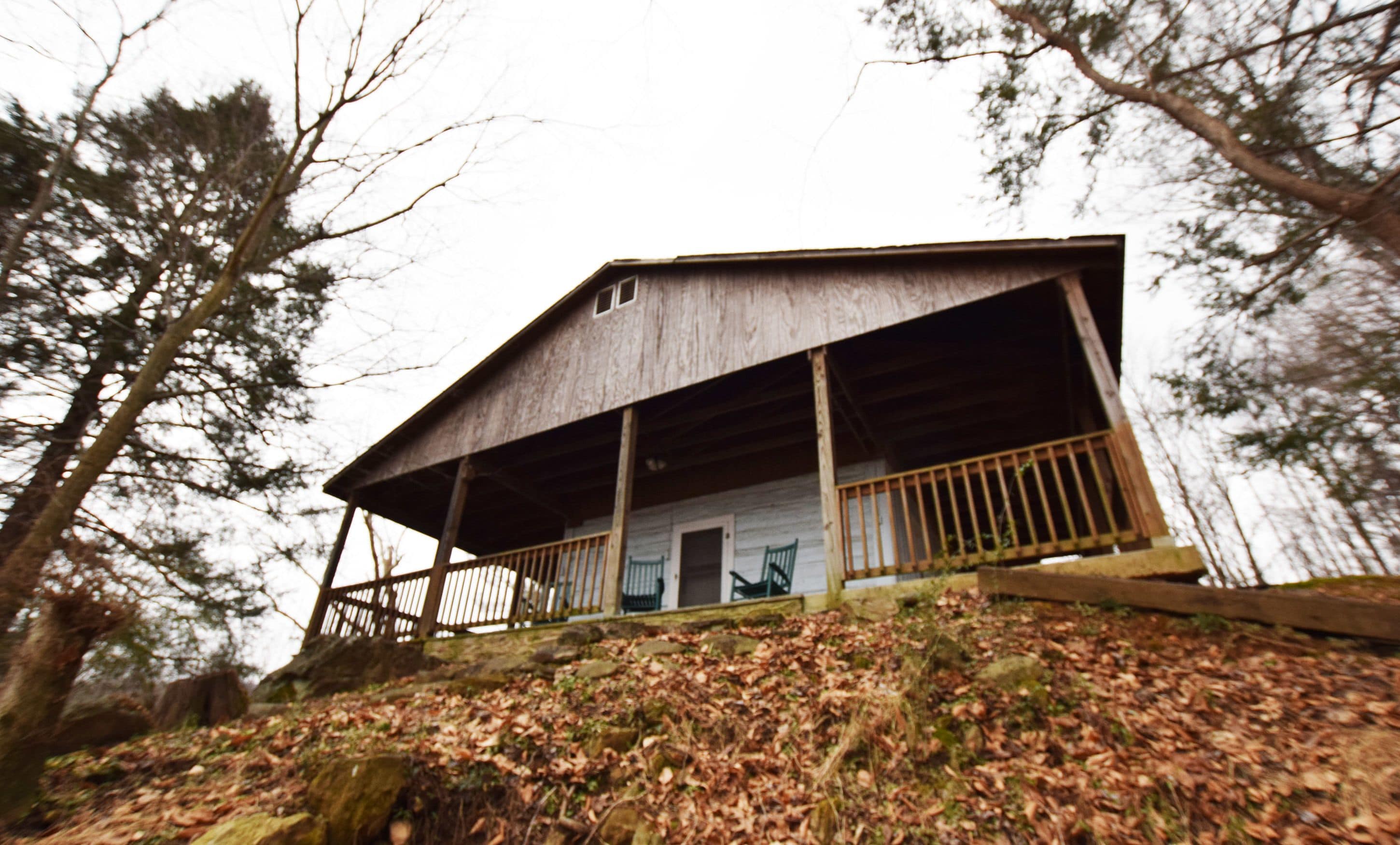 A rustic cabin with a wooden porch, surrounded by trees and a hillside covered in leaves.