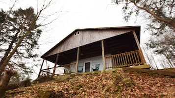 A rustic cabin with a wooden porch, surrounded by trees and a hillside covered in leaves.