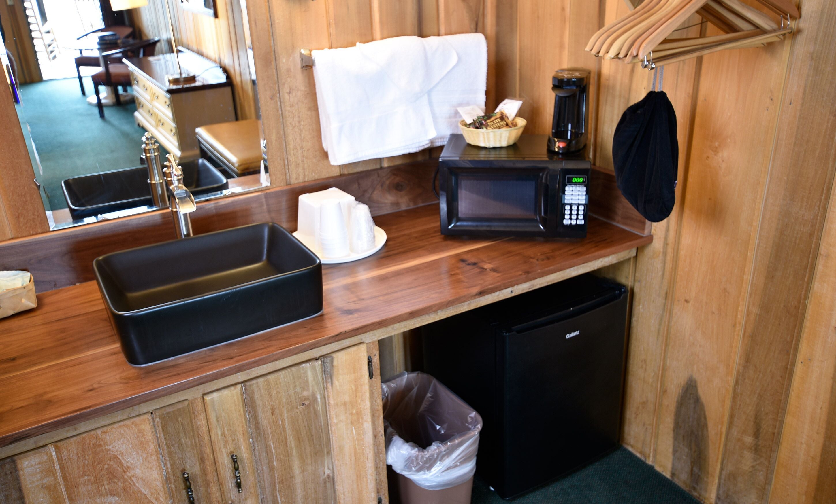Una kitchenette compacta con lavamanos, microondas, cafetera y un cubo de basura.