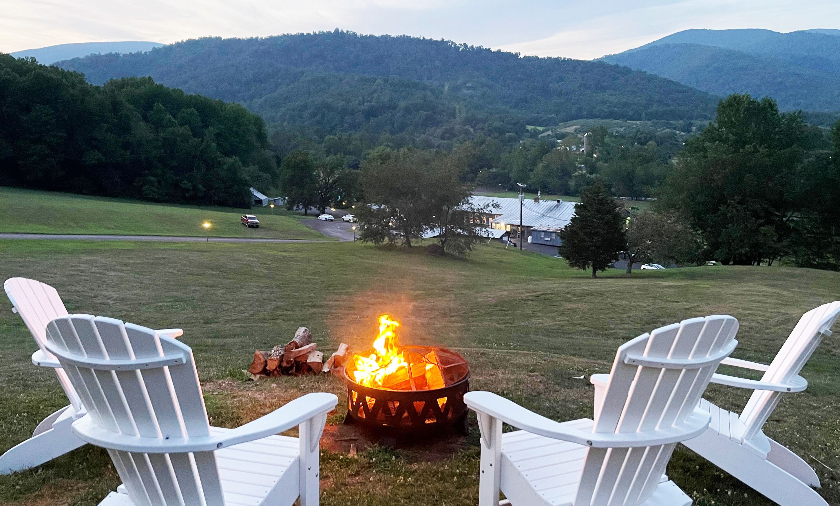 Two white Adirondack chairs facing a campfire in a grassy field with a view of a mountain range and a small building in the distance.