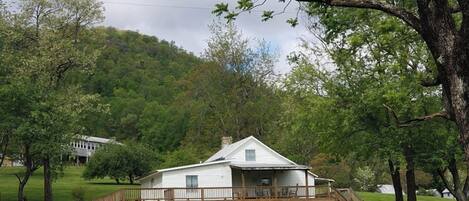 A white cabin with a wooden deck and a staircase, surrounded by lush green trees and a hill in the background.