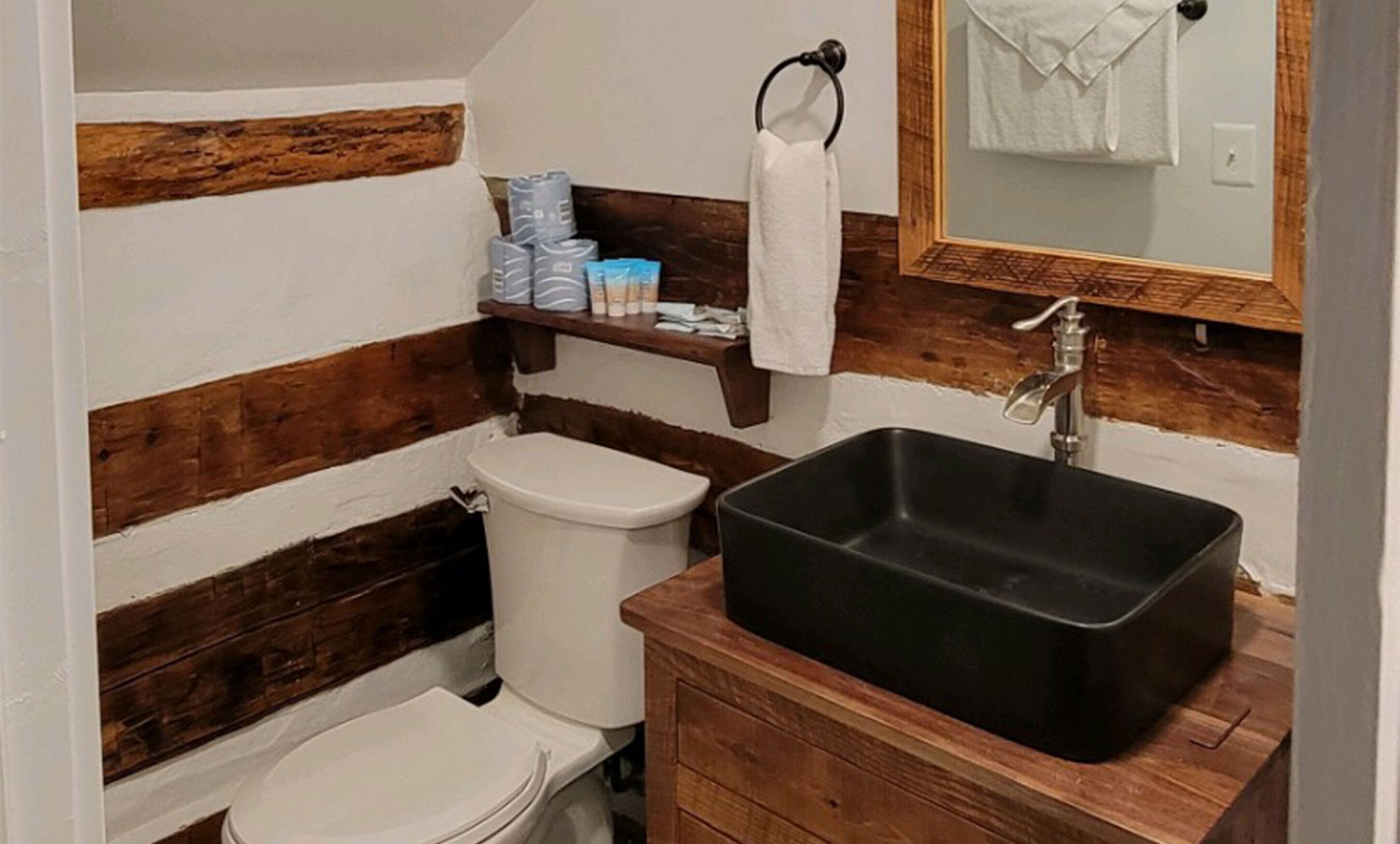 Un baño con tocador de madera, un lavamanos negro, un inodoro y un estante con artículos de tocador.