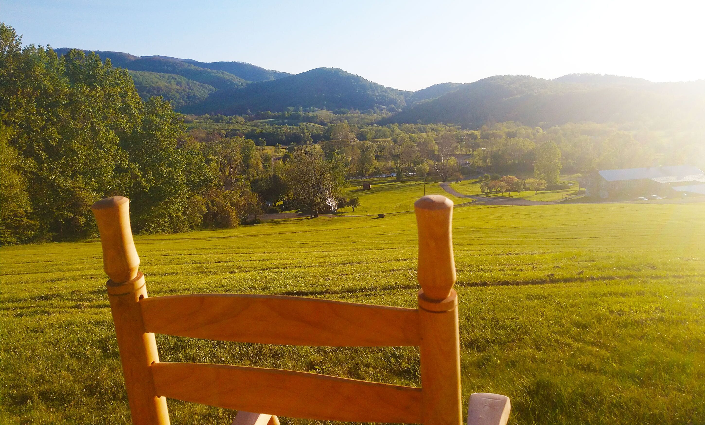 A wooden chair in a grassy field with a view of a rural landscape, including hills and trees.