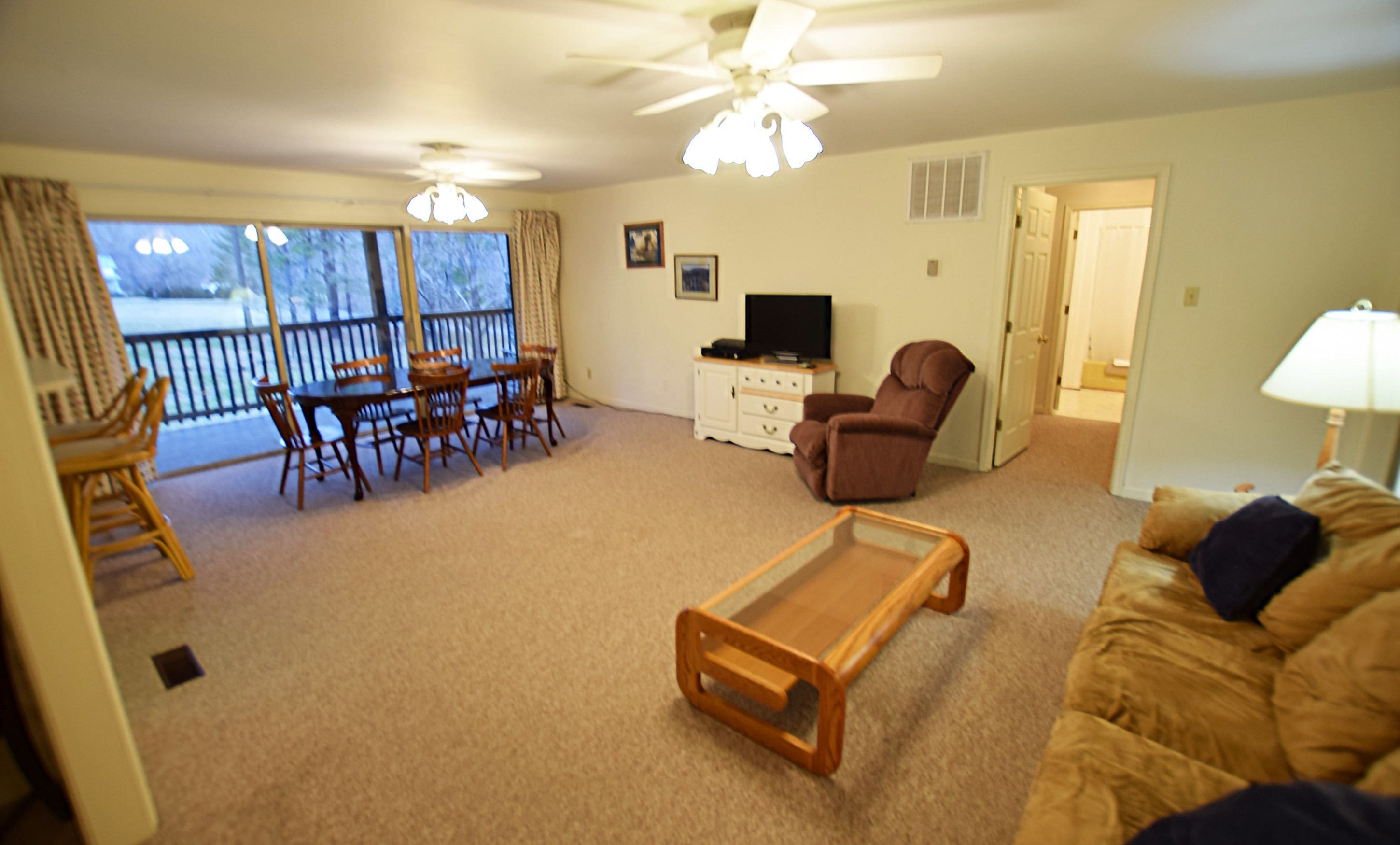 A living room with a ceiling fan, a TV on a cabinet, a brown armchair, a glass coffee table, and a sofa with cushions.
