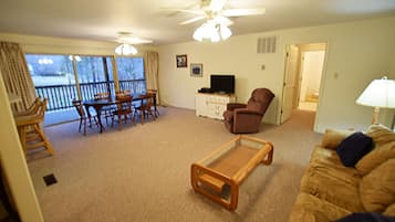 A living room with a ceiling fan, a TV on a cabinet, a brown armchair, a glass coffee table, and a sofa with cushions.