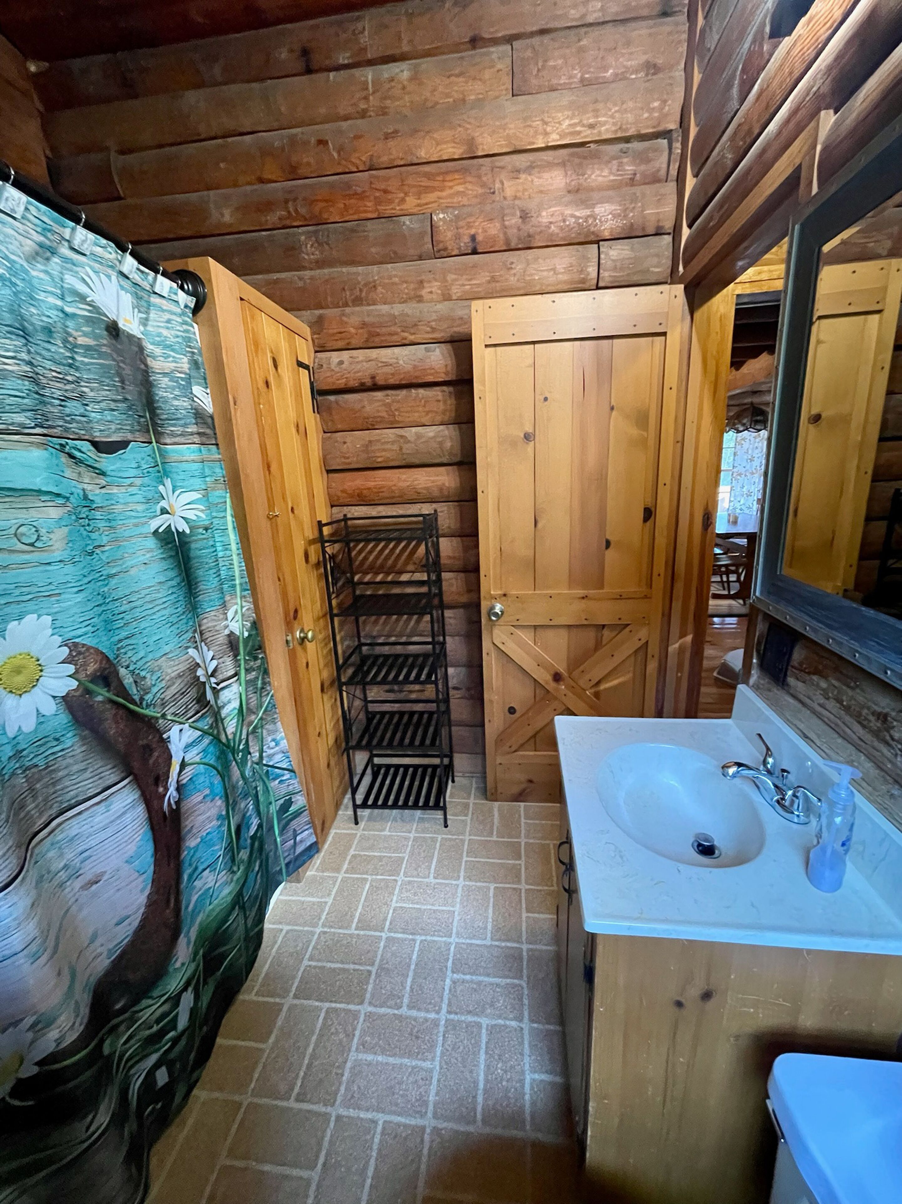 A rustic bathroom with wooden walls, a tiled floor, a wooden door, a sink with a mirror, and a shower curtain with floral patterns.