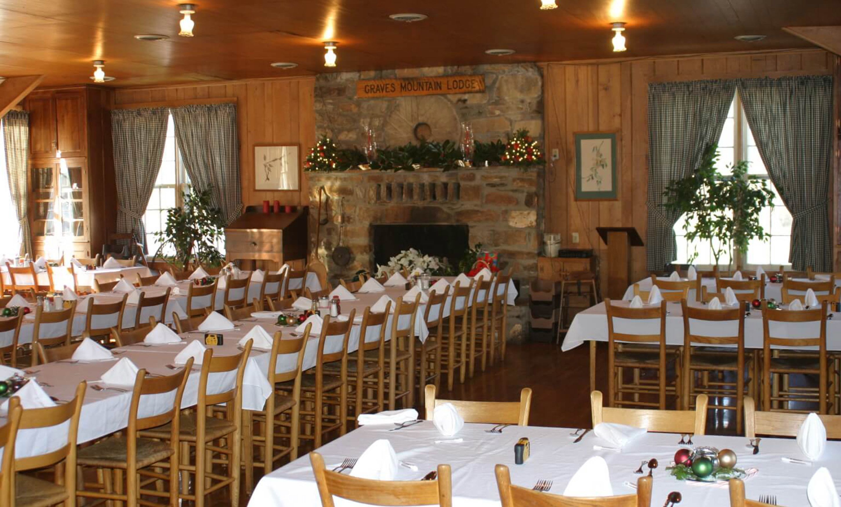 A large dining hall with multiple round tables, wooden chairs, and a stone fireplace.