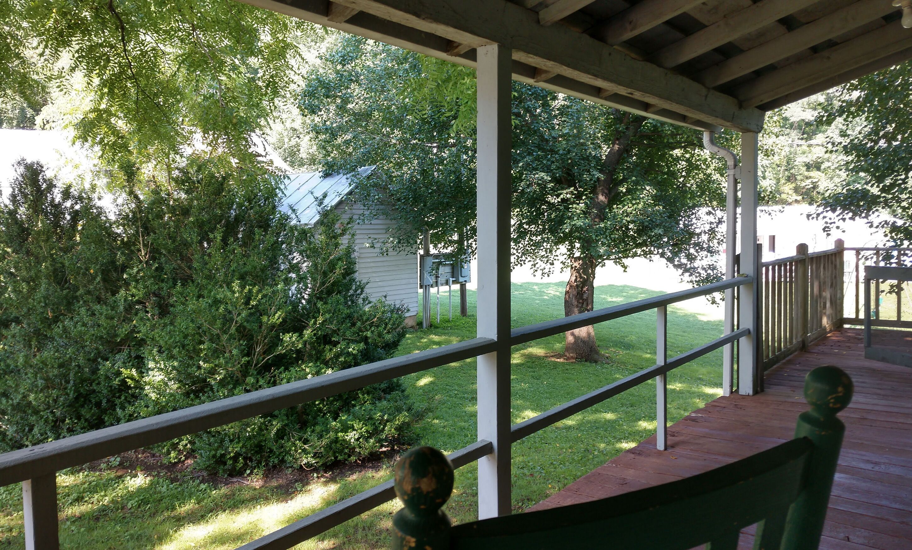 Eine überdachte Veranda mit Holzgeländer, mit Blick auf einen grünen Garten mit Bäumen und einem Haus.