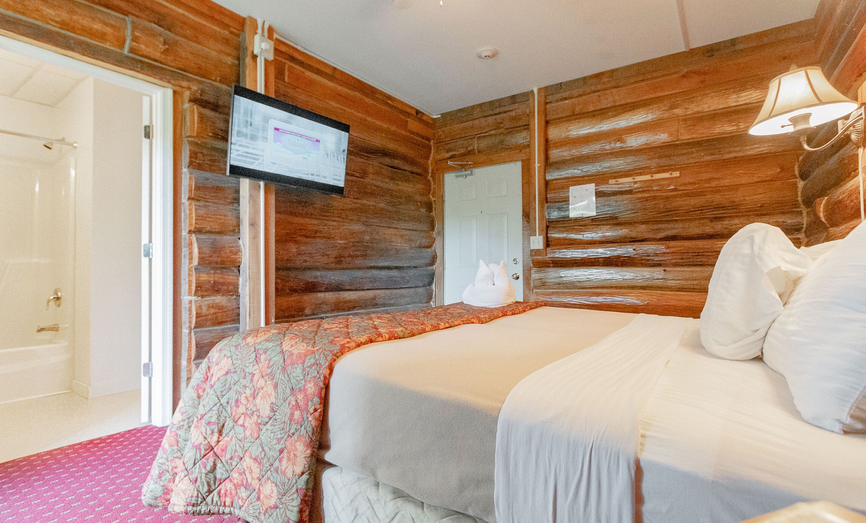Una habitación con paredes de madera, una cama con colcha estampada, un televisor en la pared y una puerta.