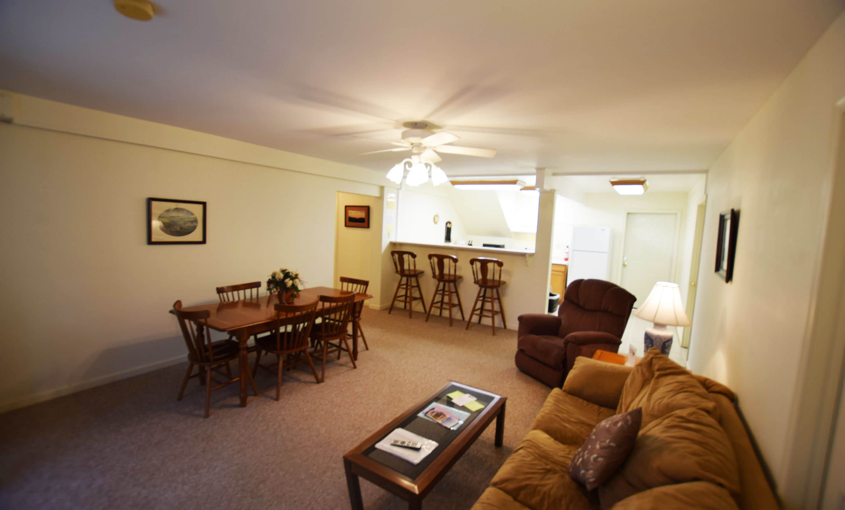A living room with a brown sofa, a coffee table, a dining table with chairs, a ceiling fan, and a kitchen area in the background.