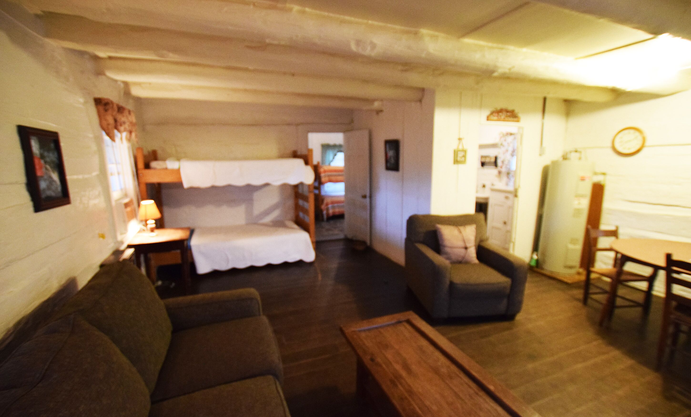 A cozy room with a bunk bed, a sofa, a small table, and a chair. There is a refrigerator and a clock on the wall.