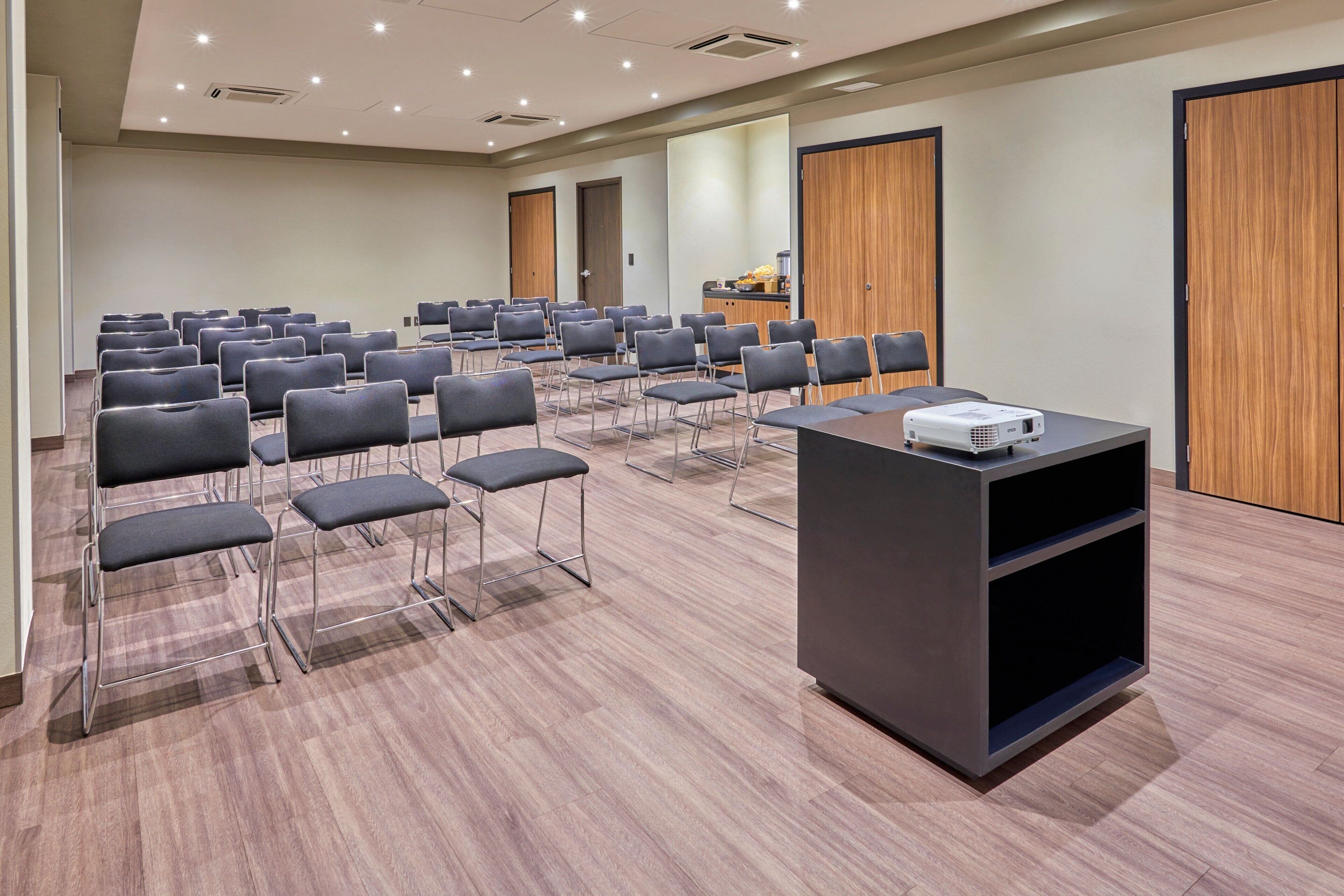 Un salón de conferencias con filas de sillas, un atril con un proyector y puertas de madera.