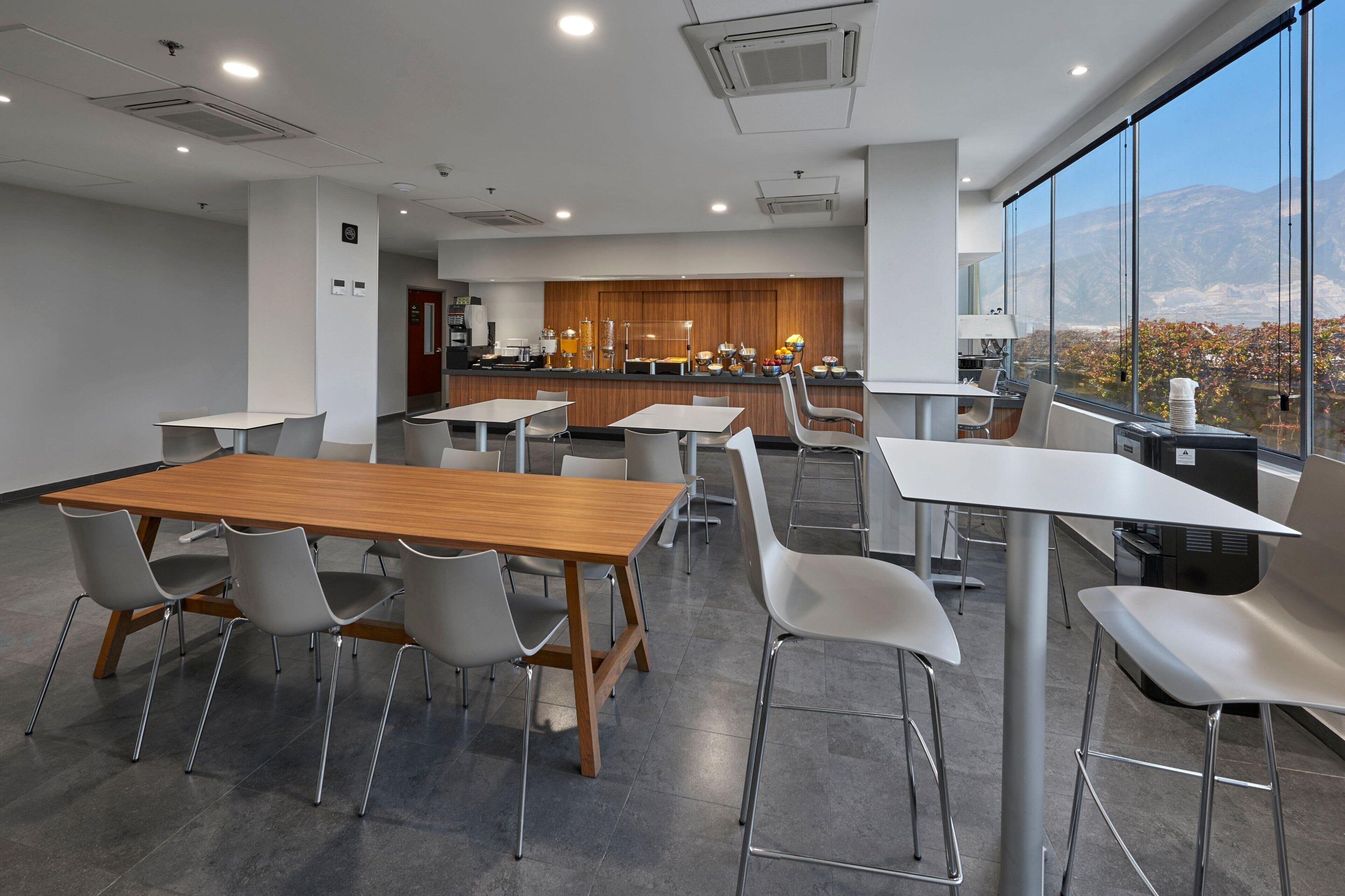 A modern dining area with a wooden table and white chairs, a kitchen in the background, and a view of mountains through large windows.
