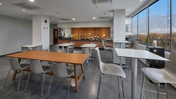 A modern dining area with a wooden table and white chairs, a kitchen in the background, and a view of mountains through large windows.