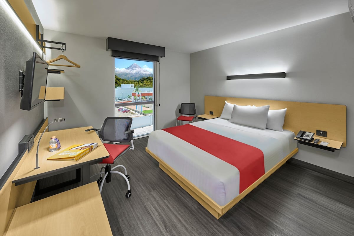 A hotel room with a bed, desk, chair, and a view of a mountain and buildings.