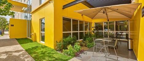 A yellow building with a patio area featuring a table and chairs, surrounded by a small garden.