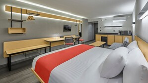 A modern hotel room with a large bed, a desk, a chair, a bench, a kitchenette, and a TV. - City Express by Marriott Tapachula (Tapachula)