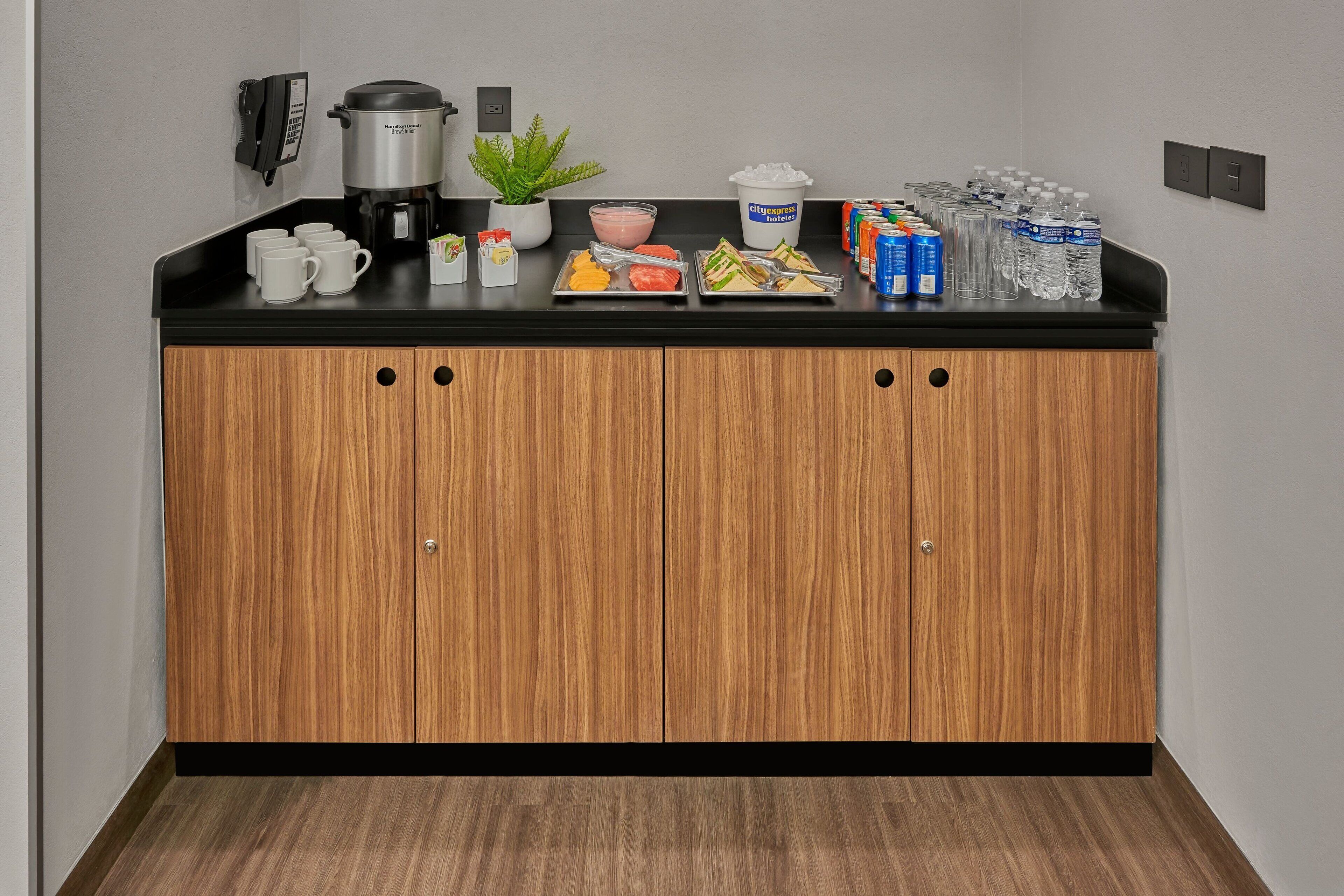 A kitchenette with a coffee maker, a bowl of fruit, a plant, and bottled water.