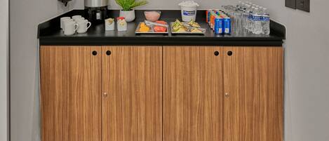 A kitchenette with a coffee maker, a bowl of fruit, a plant, and bottled water.