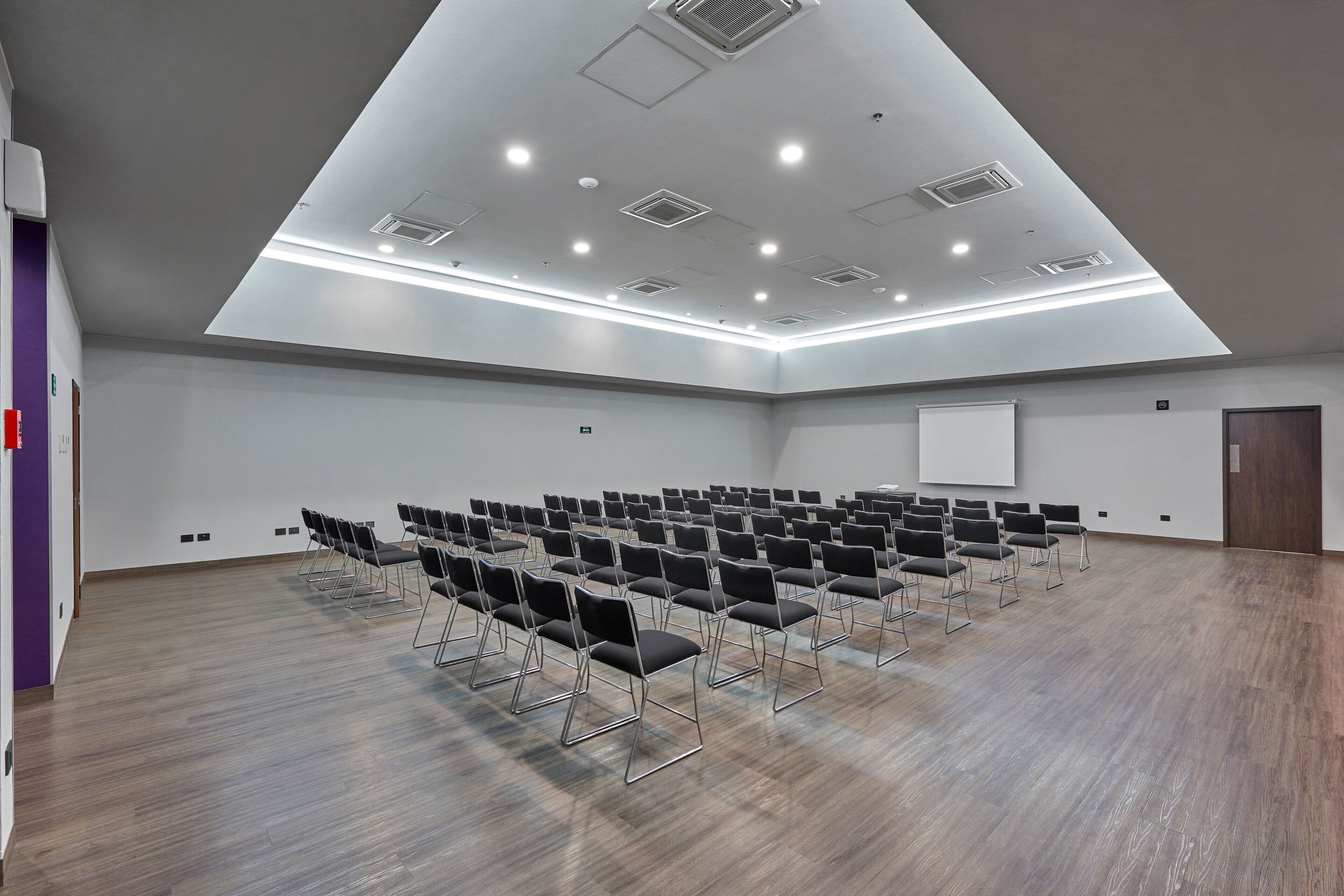 A large conference room with rows of chairs, a whiteboard, and a wooden floor.