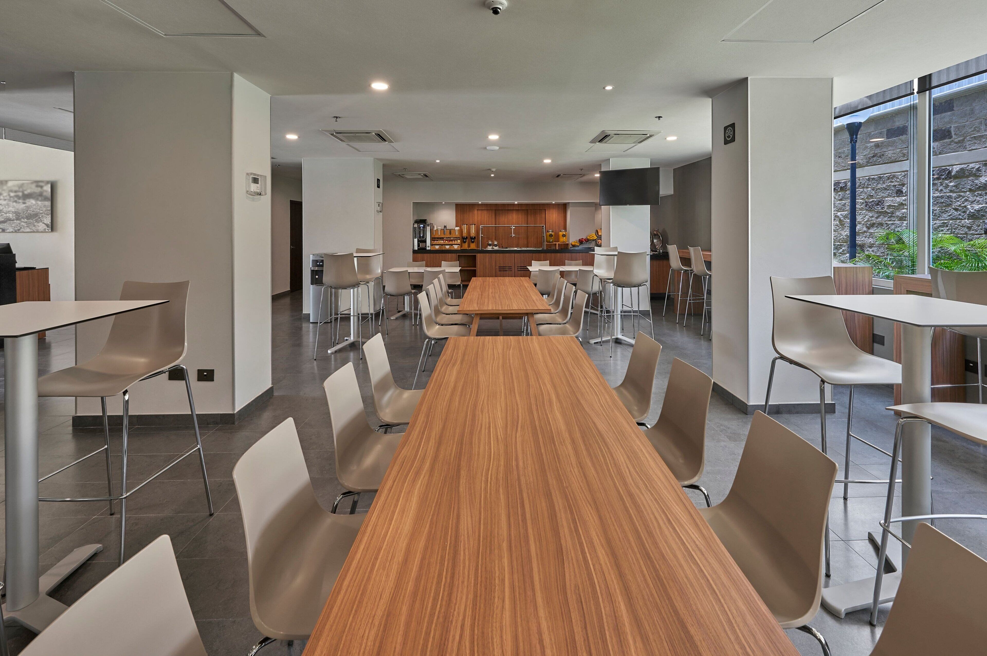 A modern conference room with a long wooden table and multiple chairs, a kitchen area in the background, and large windows providing natural light.