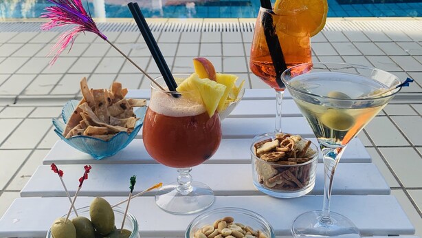 A poolside table with various drinks and snacks, a pool in the background, and a building with a tiled roof. - Hotel Deanna Golf (Cervia)
