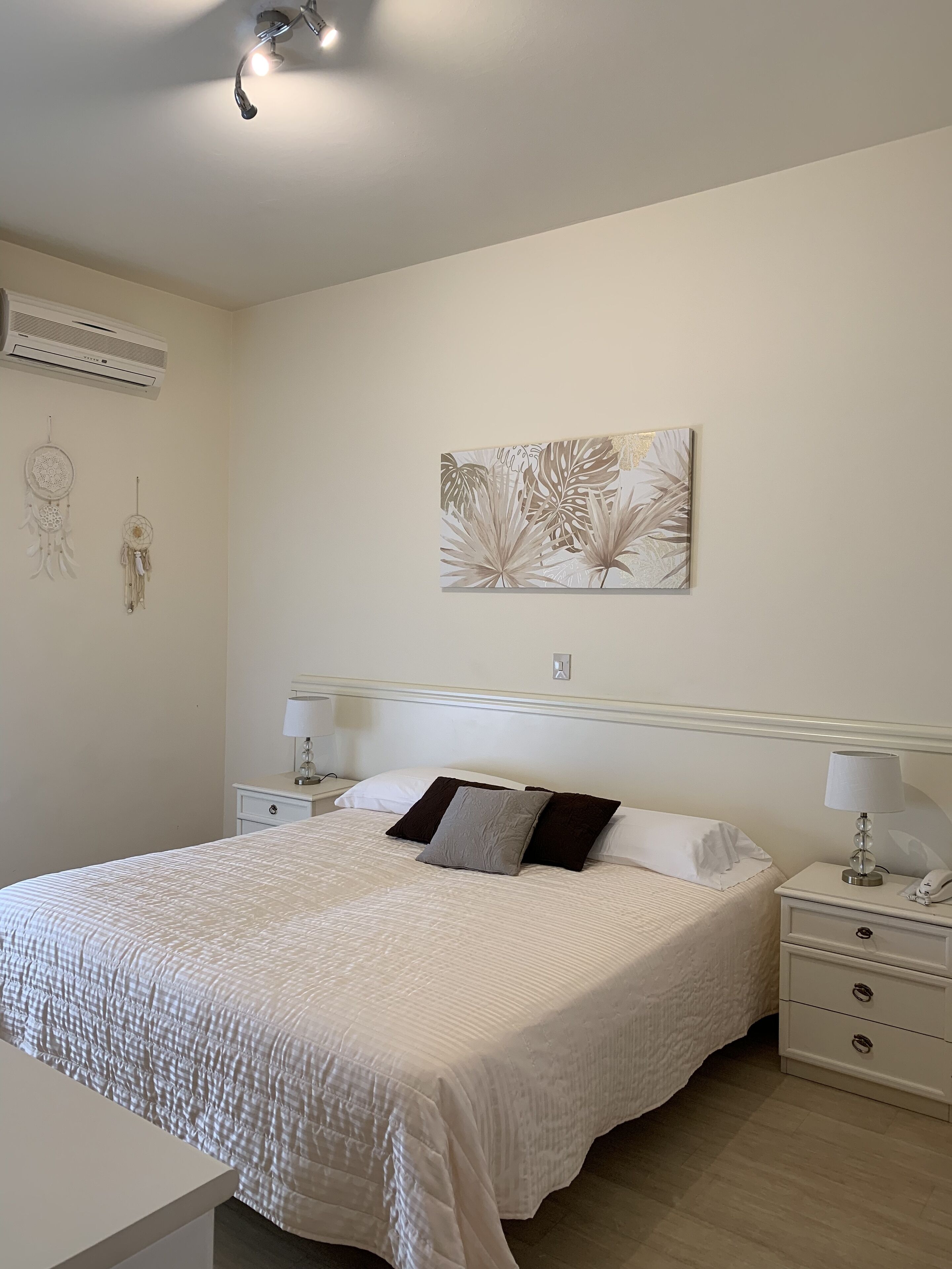 A neatly arranged bedroom with a bed, bedside tables, a wall-mounted air conditioner, and a wall art piece.