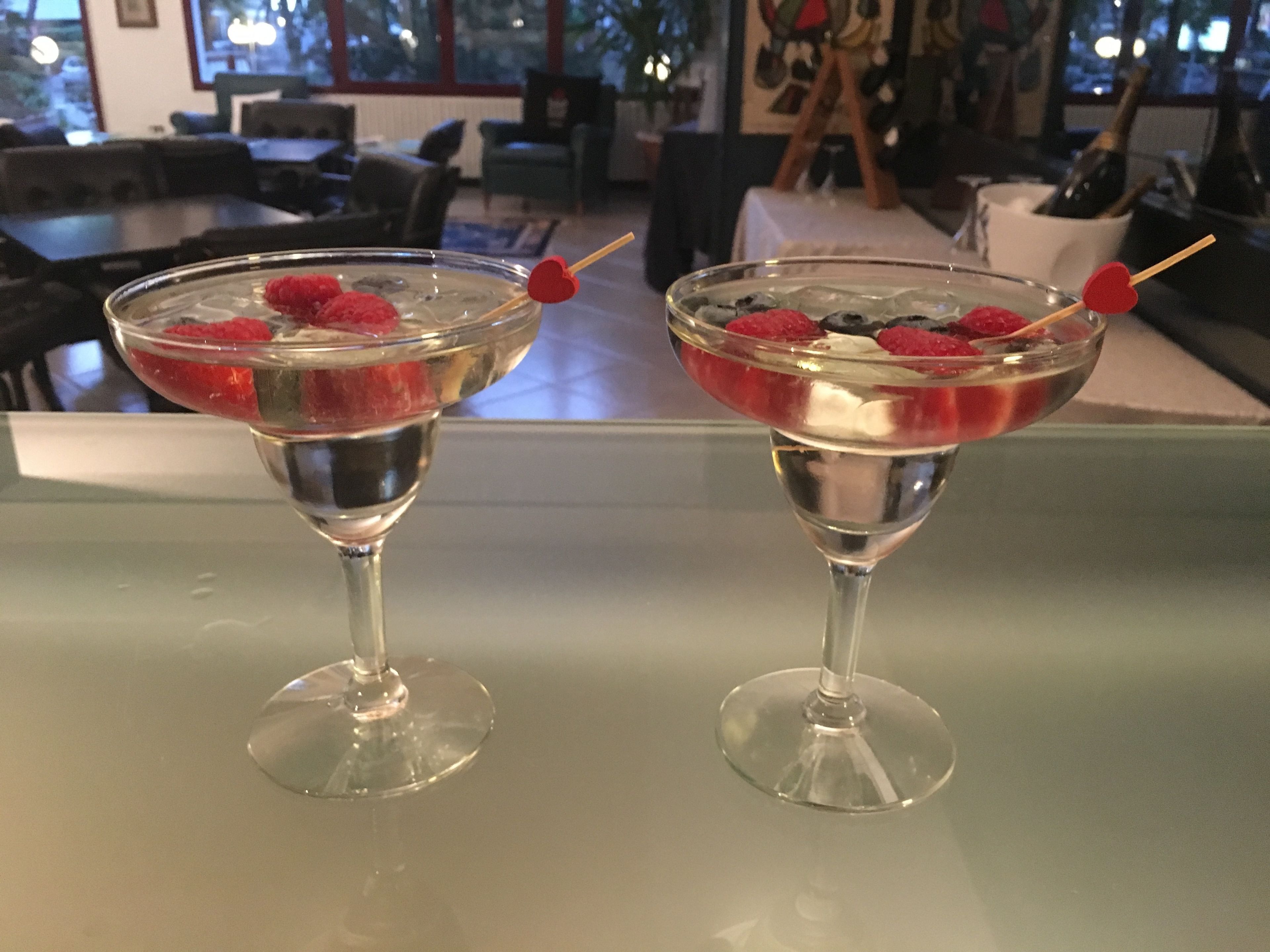 Two martini glasses with a clear liquid and raspberries on a reflective surface.
