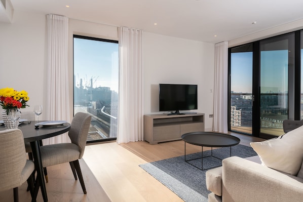A modern living room with a flat-screen TV, a dining table with chairs, a sofa, and a large window offering a city view.