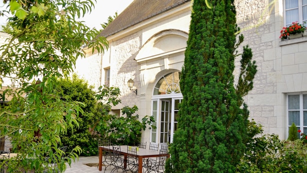 Un edificio de piedra con un patio, una mesa y sillas, y un alto árbol de coníferas.