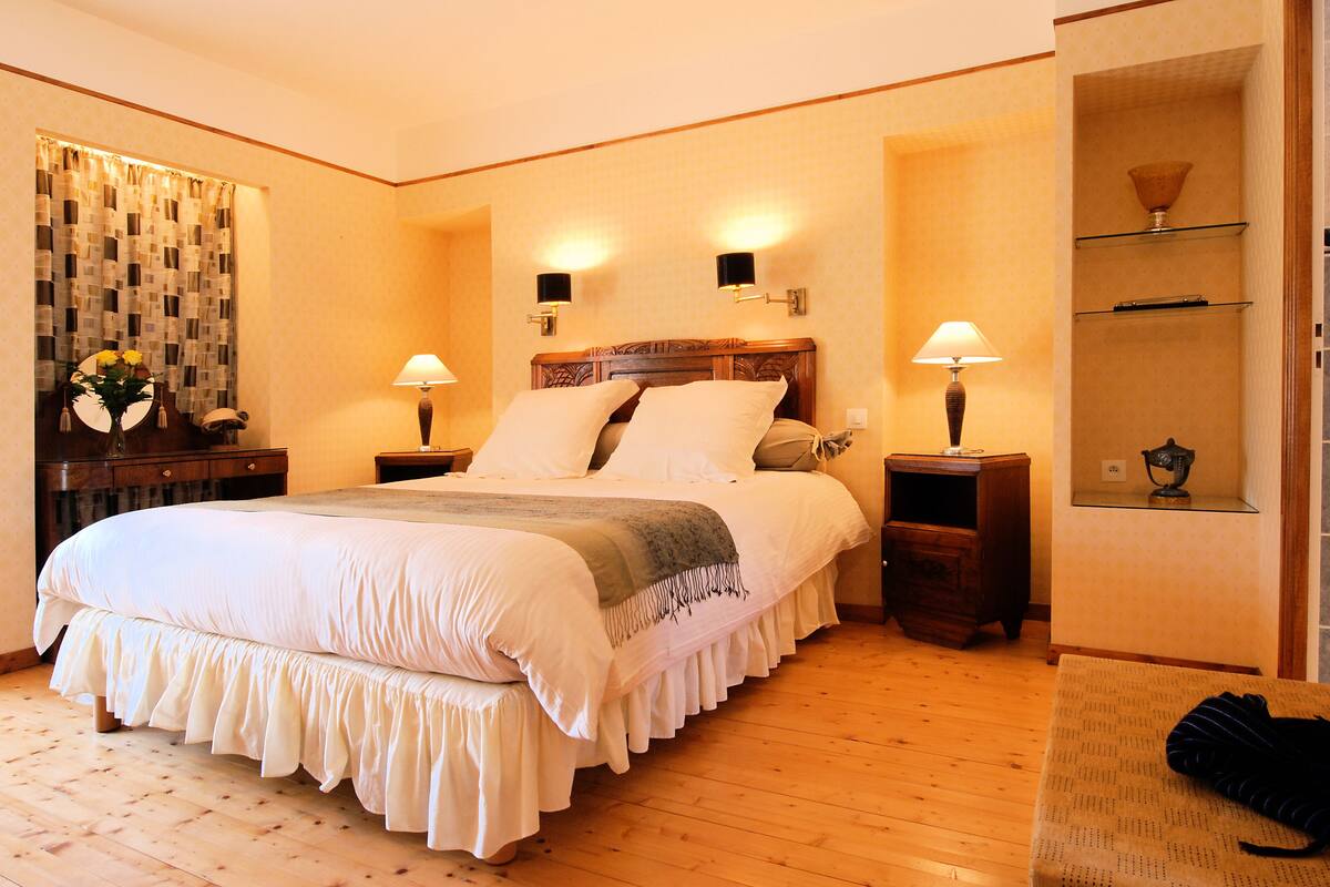 A neatly made bed with a wooden headboard, two bedside tables with lamps, a small wooden nightstand, and a wall-mounted shelf with a lamp.