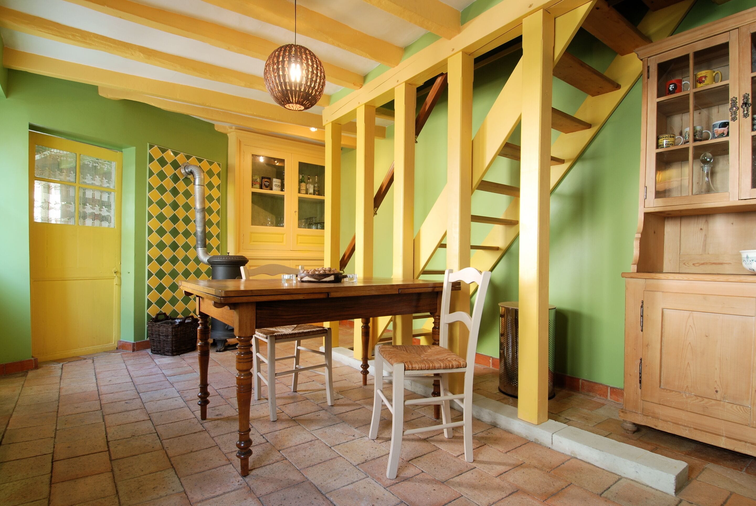 A kitchen with a wooden table, chairs, a hanging light fixture, and a staircase.