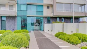 Un edificio moderno con fachada de vidrio, entrada cubierta y zona de acceso ajardinada con arbustos.