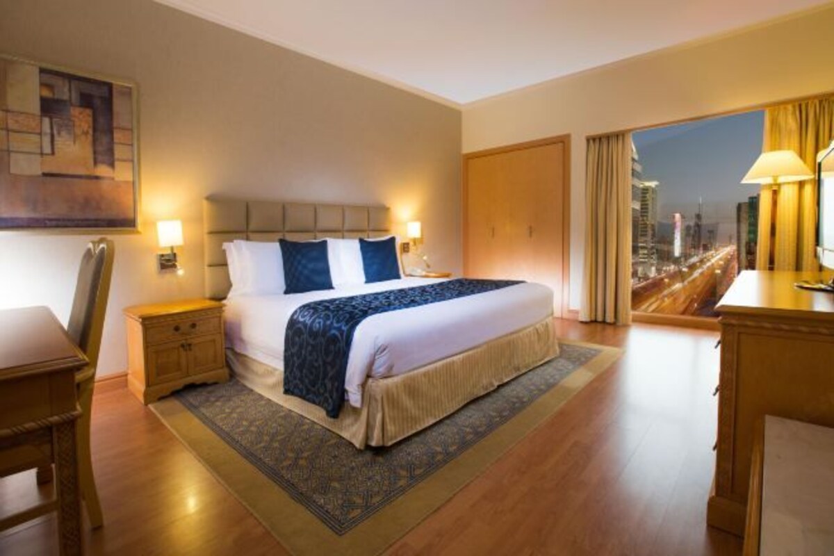Een hotelkamer met een groot bed, nachtkastjes, een bureau en uitzicht op een stadsbeeld.
