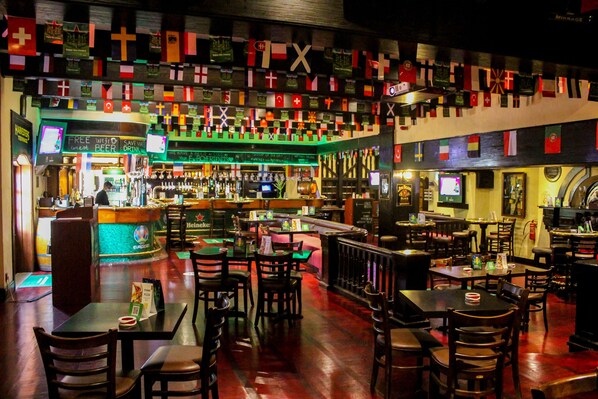 Un bar con varias banderas colgadas, mesas y sillas dispuestas para sentarse, y una barra con diferentes botellas y vasos.