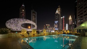 Zona de piscina nocturna con una gran estructura iluminada de caligrafía árabe, rodeada de edificios urbanos con ventanas iluminadas.