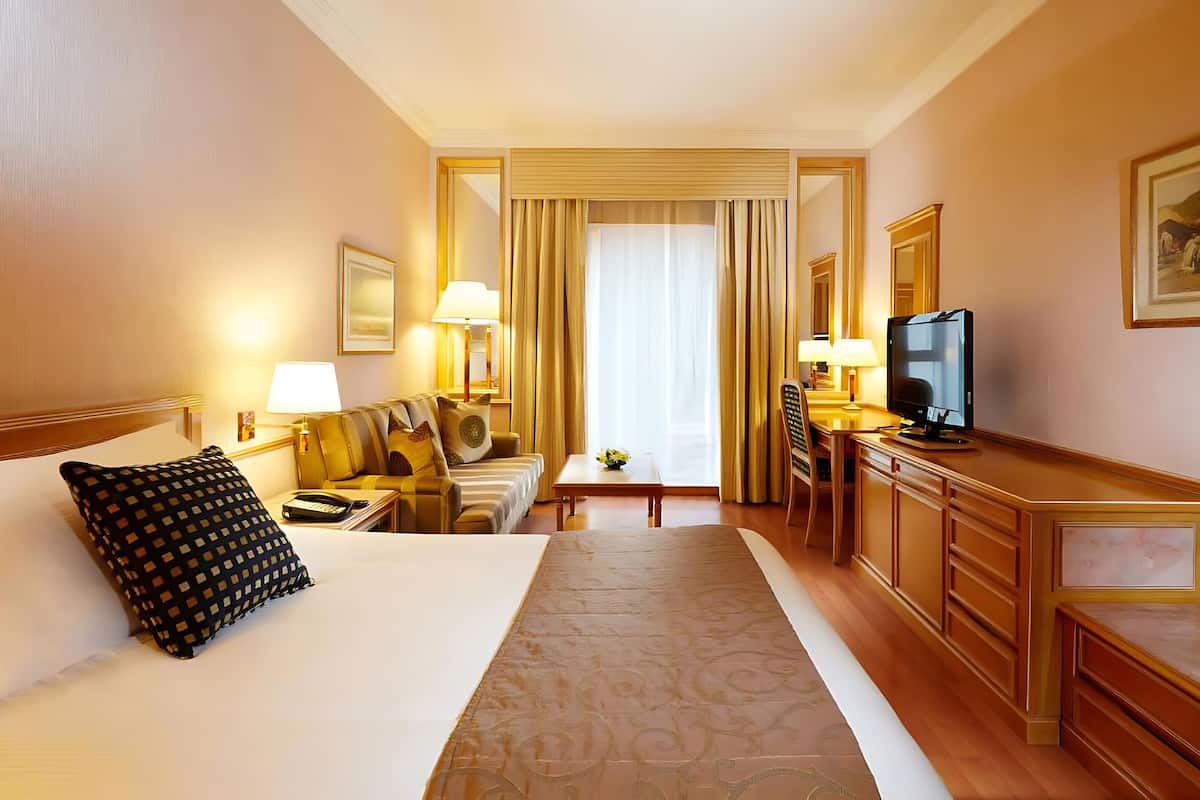 Ein Hotelzimmer mit einem großen Bett, einem Schreibtisch mit Fernseher, einem Sessel und einem kleinen Tisch mit Lampe.
