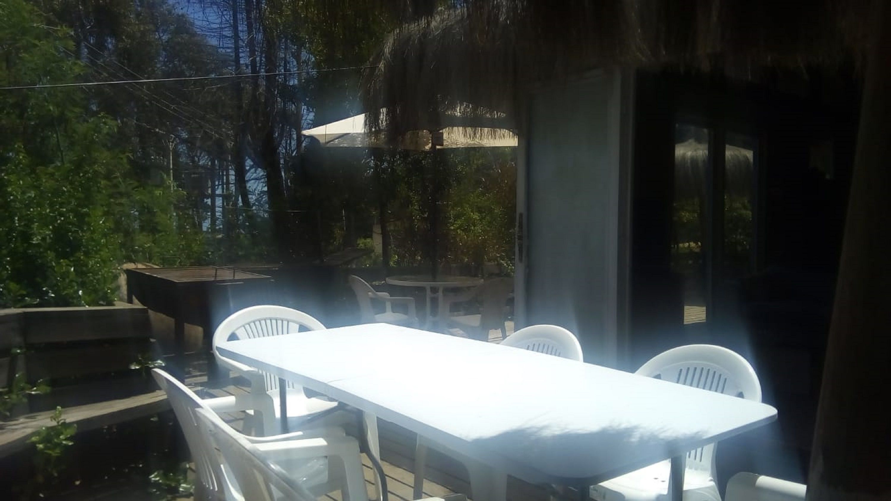 An outdoor dining area with a table and chairs, surrounded by trees and a thatched structure.