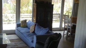 A room with a blue sofa, a black armchair, and a wooden table. There are large windows allowing natural light and views of the outdoors. - 440 Cachagua (Zapallar)