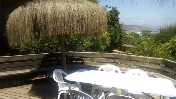 A wooden deck with a thatched umbrella, white plastic chairs, and a table. - 440 Cachagua (Zapallar)